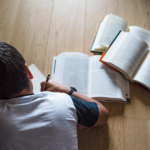 child writing while laying on wooden floor next to open books