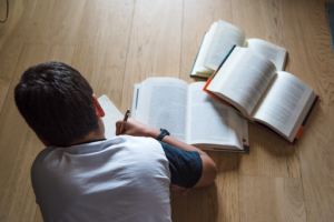 child writing while laying on wooden floor next to open books